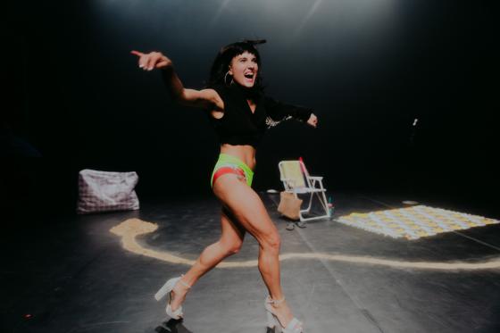 Eine Darstellerin in einer dynamischen Pose auf der Bühne, energiegeladen und ausdrucksstark. Sie trägt neonfarbene grüne Shorts und silberne High Heels, mit einem schwarzen Obertei. Im Hintergrund sind Requisiten wie eine geflochtene Tasche und ein gestreiftes Handtuch zu sehen. Eine Darstellerin in einer dynamischen Pose auf der Bühne, energiegeladen und ausdrucksstark. Sie trägt neonfarbene grüne Shorts und silberne High Heels, mit einem schwarzen Obertei. Im Hintergrund sind Requisiten wie eine geflochtene Tasche und ein gestreiftes Handtuch zu sehen.