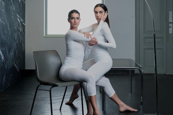 Zwei Personen in weißen Ganzkörperanzügen sitzen barfuß eng nebeneinander. Eine sitzt auf einem Stuhl, die andere halb auf ihren Beinen. Sie halten sich gegenseitig an Armen und Gesicht, während eine Stehlampe über ihnen hängt. Zwei Personen in weißen Ganzkörperanzügen sitzen barfuß eng nebeneinander. Eine sitzt auf einem Stuhl, die andere halb auf ihren Beinen. Sie halten sich gegenseitig an Armen und Gesicht, während eine Stehlampe über ihnen hängt.