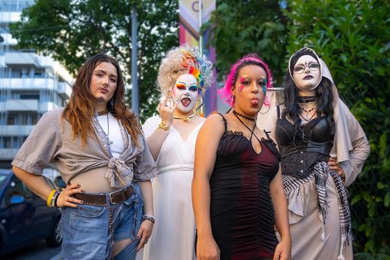 Vier Personen in auffälliger und vielfältiger Kleidung posieren selbstbewusst auf einer Straße. Eine Person trägt Jeans, geknotetes Hemd und Stiefeletten. Eine zweite Person trägt ein langes weißes Kleid und bunte Perücke mit weiß geschminktem Gesicht. Die dritte Person trägt ein kurzes schwarzes Kleid mit Netzstrumpfhose und pinken Haaren. Die vierte Person trägt ein Korsett mit langer Robe und Gesichtsschminke im Stil einer Nonne. Im Hintergrund sind Bäume, ein Auto und Gebäude zu sehen. Vier Personen in auffälliger und vielfältiger Kleidung posieren selbstbewusst auf einer Straße. Eine Person trägt Jeans, geknotetes Hemd und Stiefeletten. Eine zweite Person trägt ein langes weißes Kleid und bunte Perücke mit weiß geschminktem Gesicht. Die dritte Person trägt ein kurzes schwarzes Kleid mit Netzstrumpfhose und pinken Haaren. Die vierte Person trägt ein Korsett mit langer Robe und Gesichtsschminke im Stil einer Nonne. Im Hintergrund sind Bäume, ein Auto und Gebäude zu sehen.