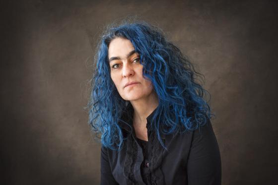 Porträt einer Frau mit langen, lockigen, blauen Haaren, die in einem dunklen Oberteil vor neutralem Hintergrund sitzt und direkt in die Kamera blickt.“ Porträt einer Frau mit langen, lockigen, blauen Haaren, die in einem dunklen Oberteil vor neutralem Hintergrund sitzt und direkt in die Kamera blickt.“