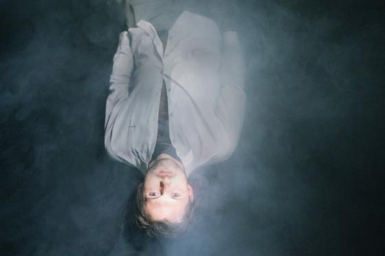 Eine Person liegt in dichter Nebel- oder Rauchkulisse. Sie trägt ein helles Hemd und blickt nach oben, während sie von oben beleuchtet wird. Eine Person liegt in dichter Nebel- oder Rauchkulisse. Sie trägt ein helles Hemd und blickt nach oben, während sie von oben beleuchtet wird.